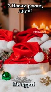 Новорічна листівка для друзів із кількома червоними шапками Санти на пухнастій білій поверхні перед каміном, оточеними гірляндою й новорічними прикрасами.