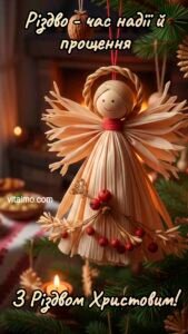 Різдвяне привітання із солом’яним янголом‑іграшкою з червоними ягідками, що висить на зеленій ялинці біля запалених свічок.